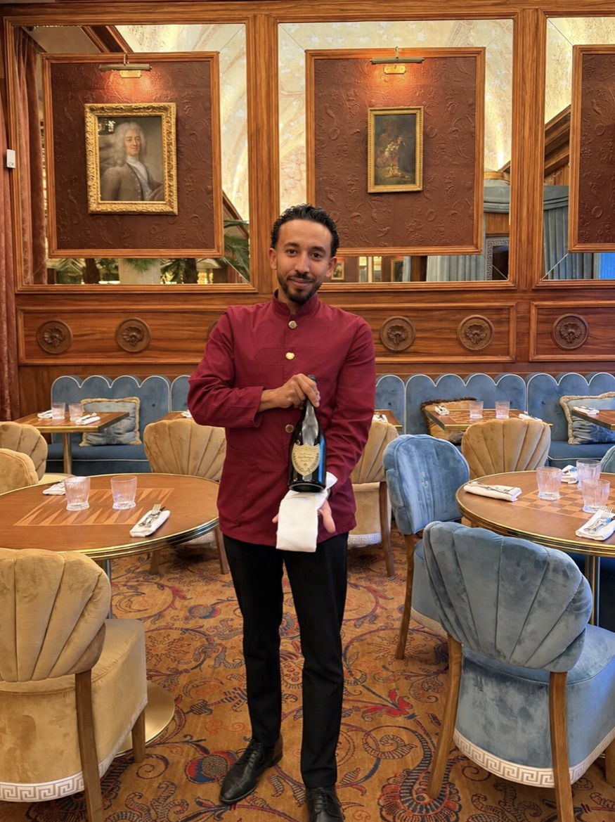 Ryan en veste bordeaux, une bouteille de champagne à la main, dans la salle boisée d'une grande maison parisienne